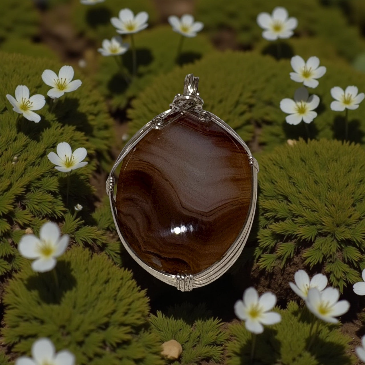 Brown stone pendant with silver frame on green foliage and white flowers