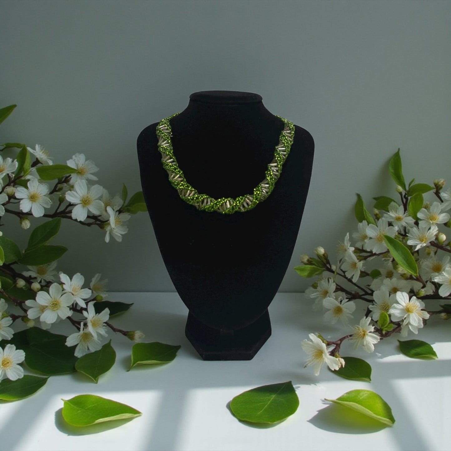 Green necklace on a black mannequin bust with white flowers and green leaves in the background