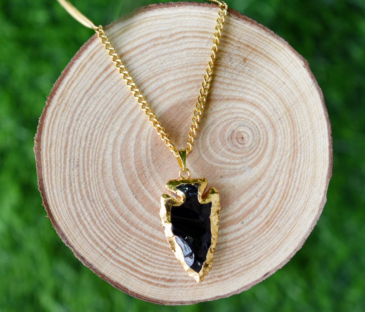Gold necklace with a black pendant on a wooden disc against a green grass background