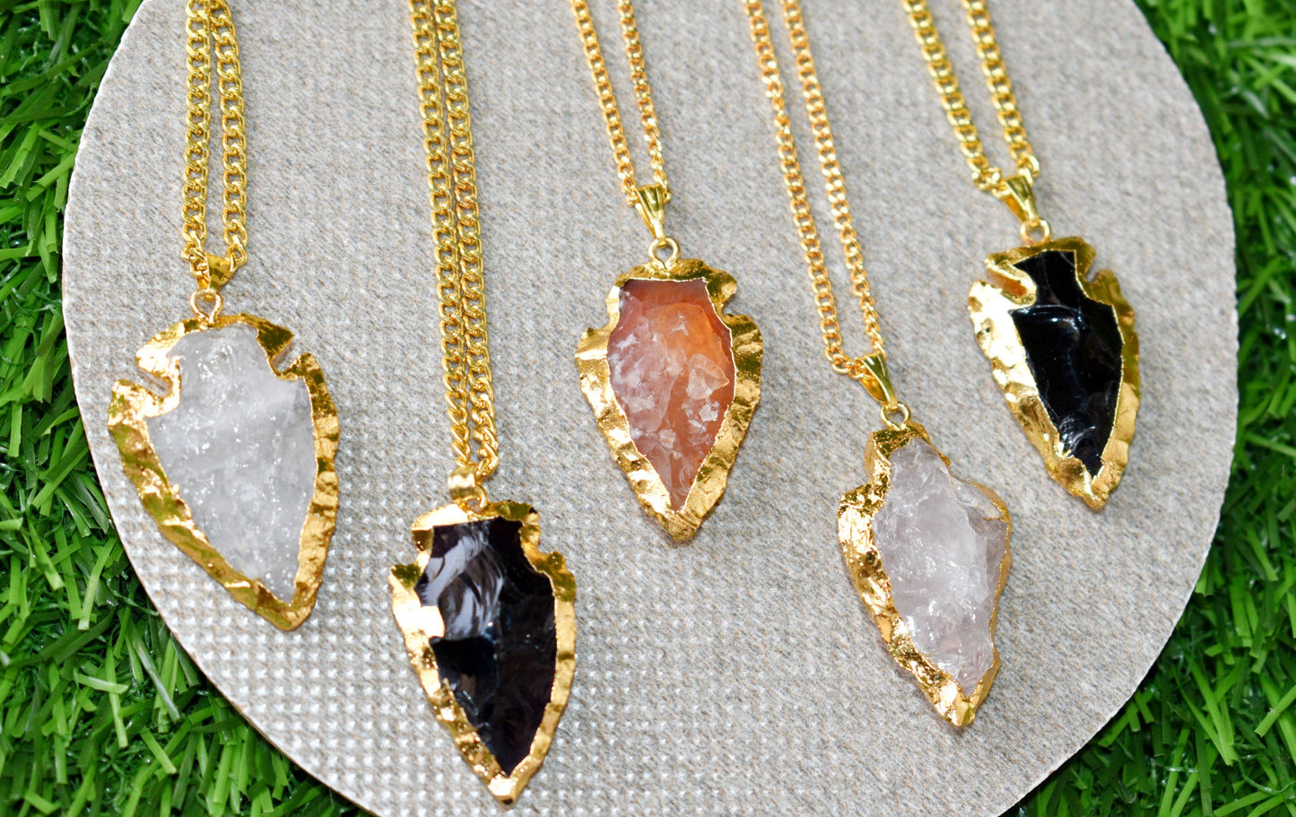 Five gold necklaces with different stone pendants on a gray stand against a grass background