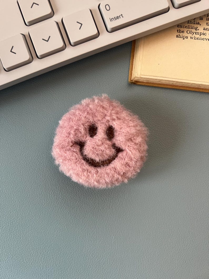 Pink fuzzy smiley face object on a desk with a keyboard and paper in the background