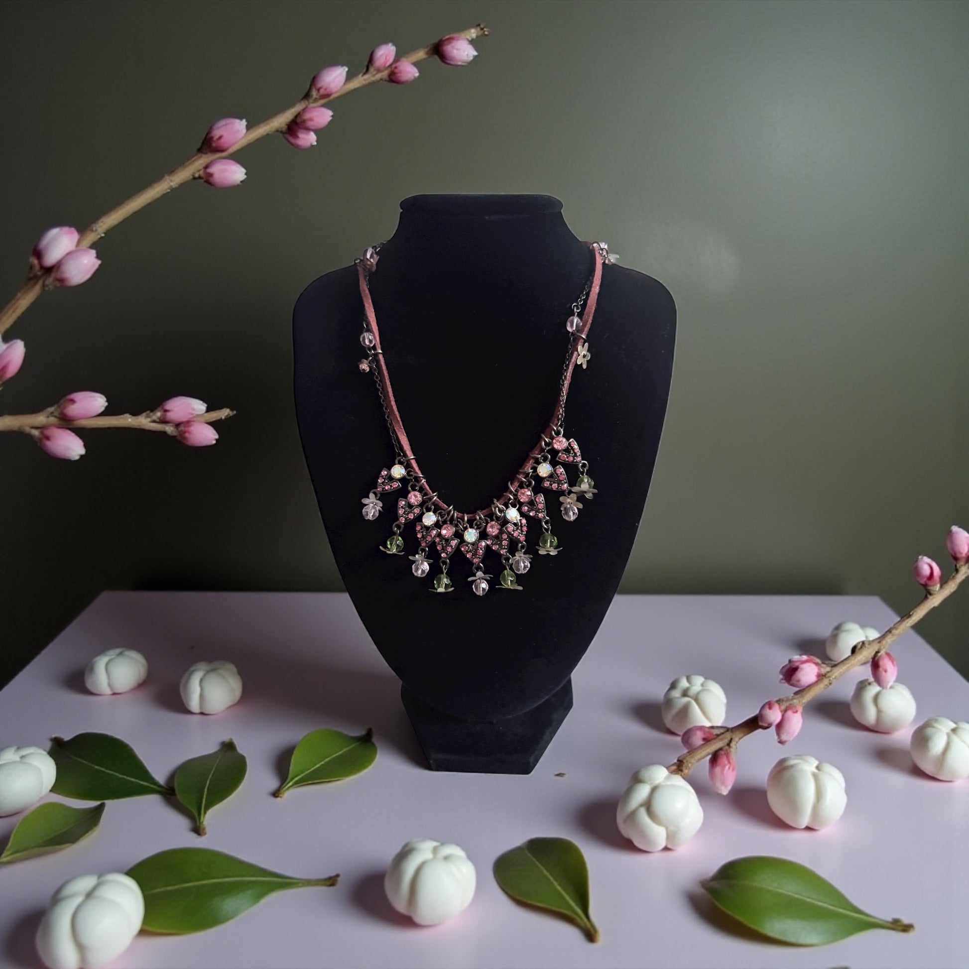 Necklace on a black mannequin bust with decorative branches and white fruits on a light pink surface.