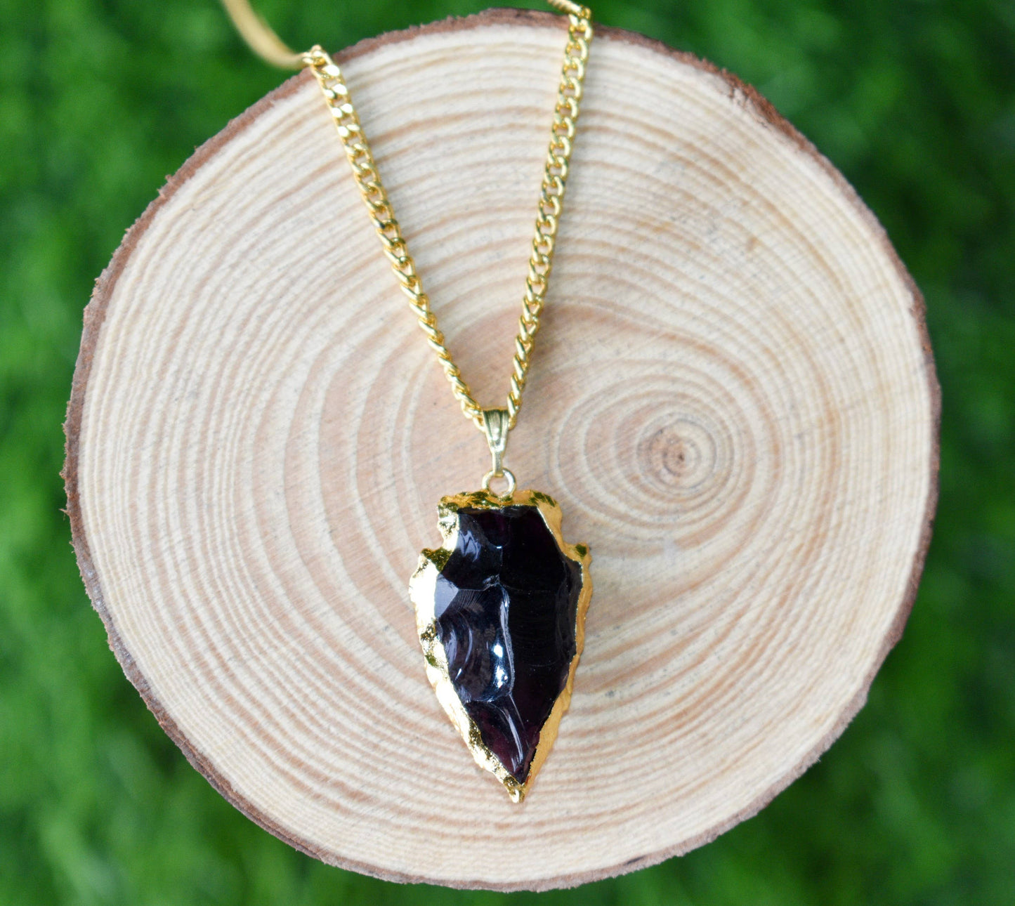 Gold necklace with a black pendant on a wooden slice against a green grass background